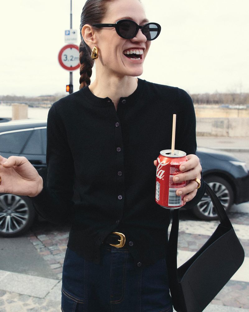 Woman smiles outdoors, holding a Coca-Cola can with a straw, wearing a black cardigan, blue jeans, and gold jewelry on a city street.