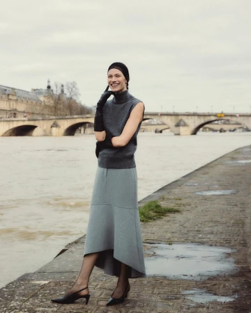 A smiling woman in a sleeveless sweater and skirt stands by the Seine River in Paris, with the Pont Neuf and buildings in the background.