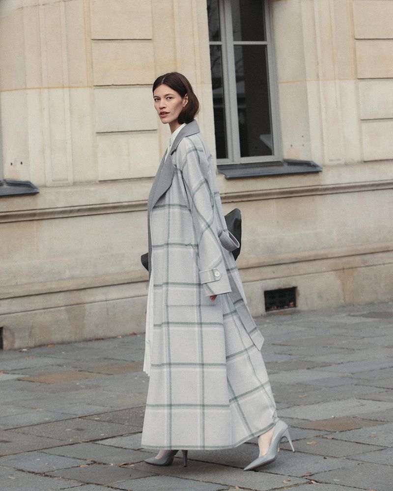 A woman in a grey plaid coat walks on a city street, complemented by grey heels, with a neutral building facade in the background.