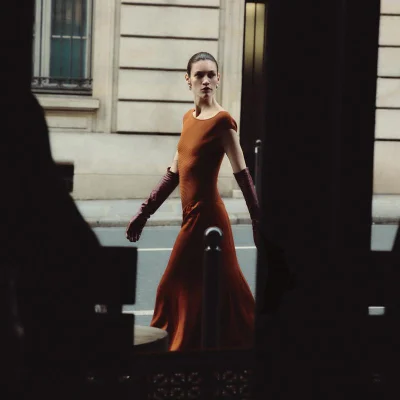 Model wearing a rust-colored dress and long burgundy gloves walking on a city street.