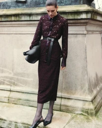 Model in burgundy lace top and matching skirt wearing a thick black belt and holding a black handbag outdoors