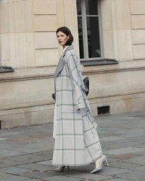 Model wearing a long grey plaid coat and heels walking past a classic building exterior