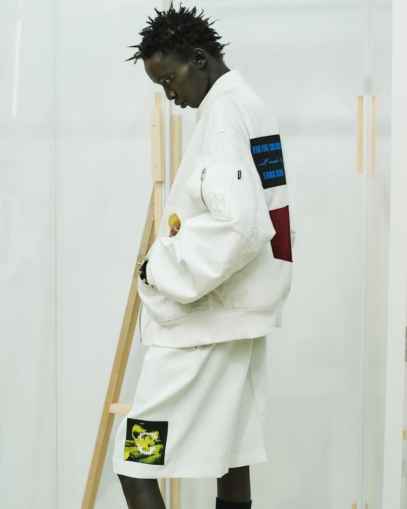 Model wearing a white graphic bomber jacket and matching shorts standing in profile next to a wooden studio frame.