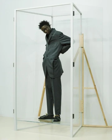 Stylish adult Black man in a grey suit and spiked shoes poses in a transparent box against a white wall in a minimalist studio setting.