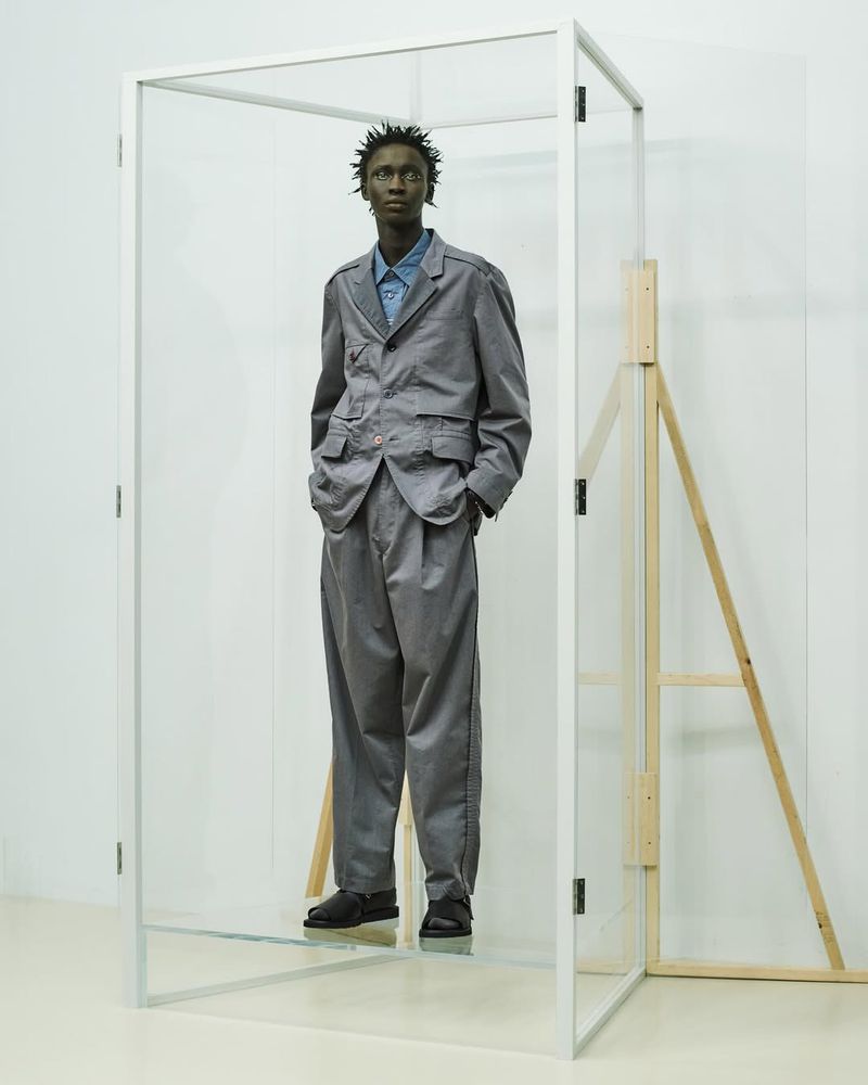 An adult male model wearing a gray suit and blue shirt poses in a clear glass cube against a white wall with a wooden structure behind.