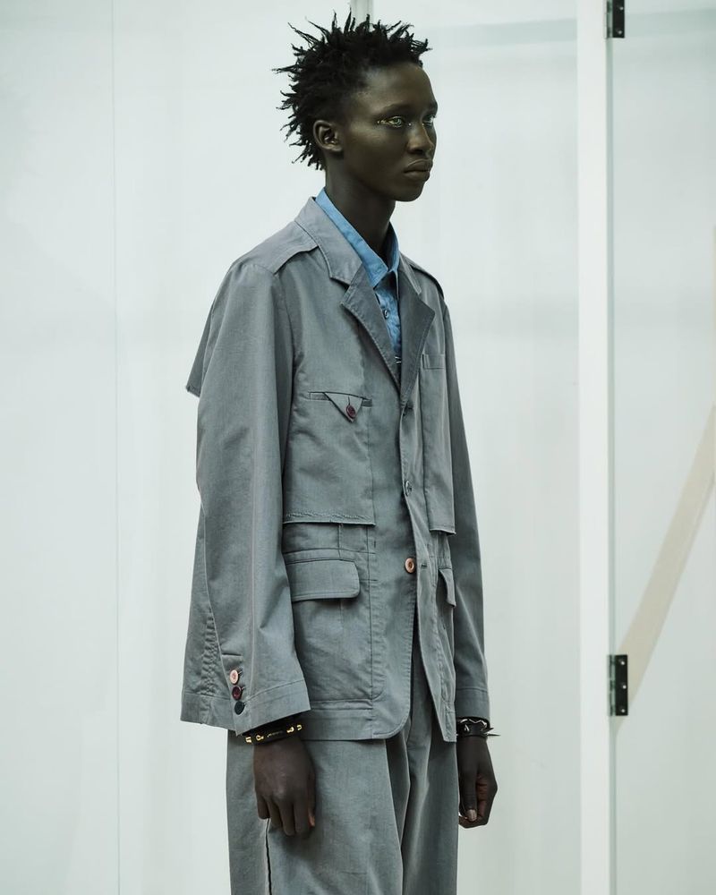 Male model in a grey jacket and pants standing against a white background, showcasing fashion attire with a serious facial expression.