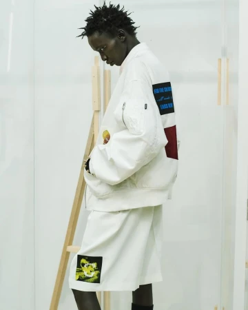Model wearing a white graphic bomber jacket and matching shorts standing in profile next to a wooden studio frame.