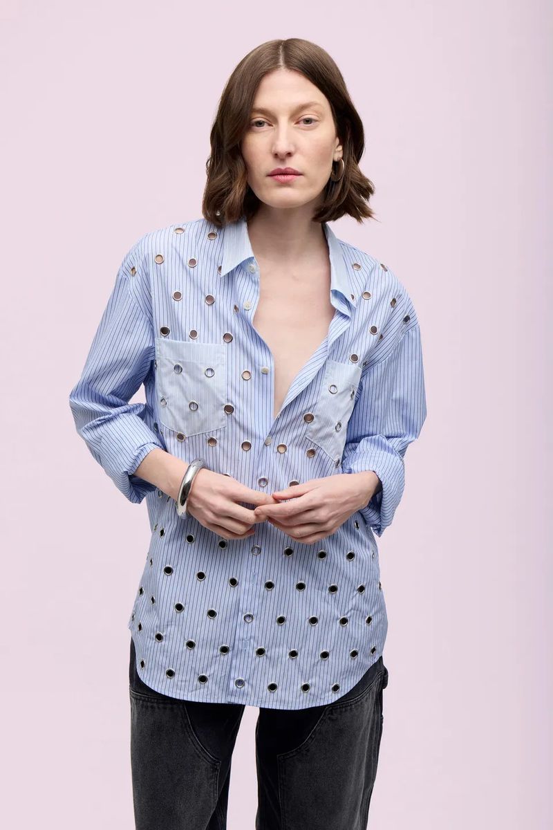 Adult woman posing indoors with neutral expression, wearing a pinstriped eyelet shirt and dark-wash jeans in a fashion studio shot.