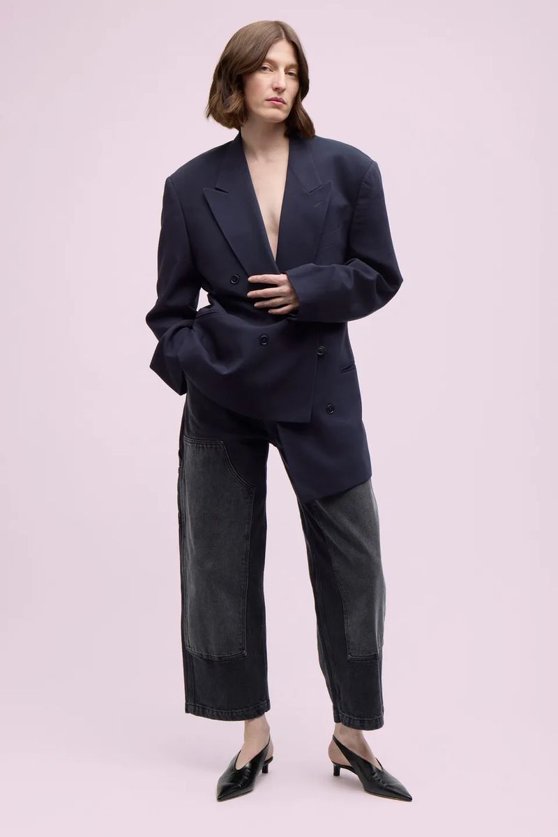 A woman in an oversized navy blazer and patchwork jeans stands in front of a soft pink background. She is wearing black heels.