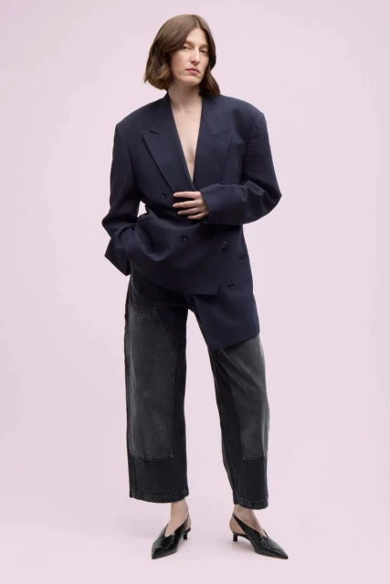 A woman in an oversized navy blazer and patchwork jeans stands in front of a soft pink background. She is wearing black heels.