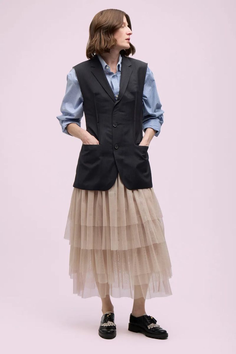 Full-length shot of a woman in a blue button-down, black vest, and layered tulle skirt, standing against a light pink backdrop in a studio setting.