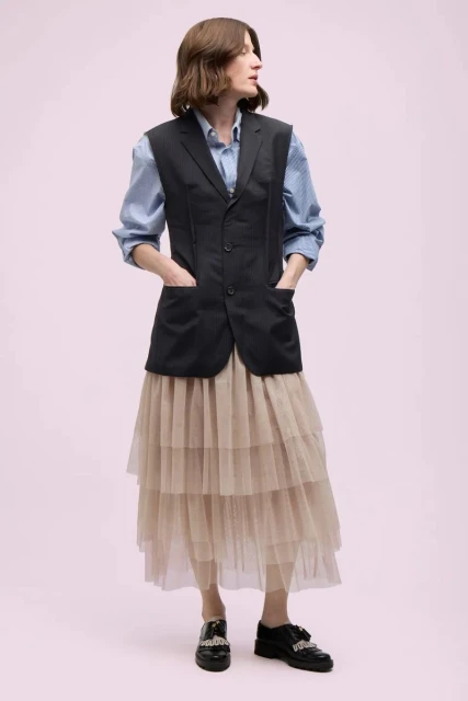 Full-length shot of a woman in a blue button-down, black vest, and layered tulle skirt, standing against a light pink backdrop in a studio setting.
