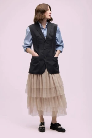 Full-length shot of a woman in a blue button-down, black vest, and layered tulle skirt, standing against a light pink backdrop in a studio setting.