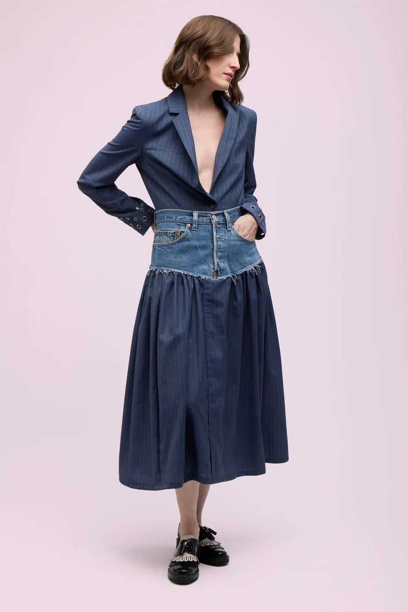 Full-length shot of a woman in a blue blazer and deconstructed denim skirt, posing in front of a pink backdrop in a fashion editorial.