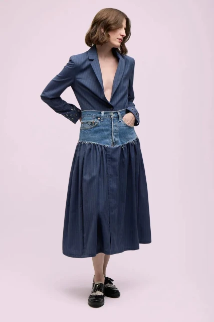 Full-length shot of a woman in a blue blazer and deconstructed denim skirt, posing in front of a pink backdrop in a fashion editorial.
