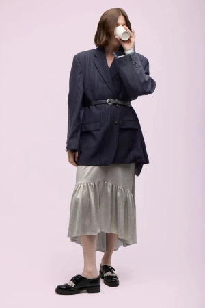 Woman in oversized pinstripe blazer and skirt drinks from paper cup against pink backdrop in a studio shot, showcasing fashion.