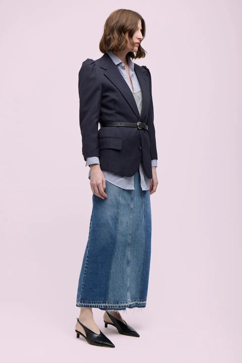 A woman poses in a pinstripe blazer, blue button-down shirt, and a patchwork denim skirt against a light pink backdrop in a fashion editorial.