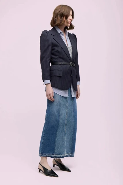 A woman poses in a pinstripe blazer, blue button-down shirt, and a patchwork denim skirt against a light pink backdrop in a fashion editorial.
