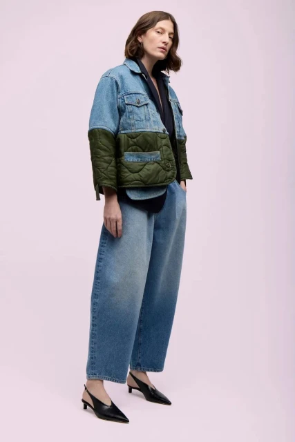 Adult female posing in studio wearing a denim jacket with quilted sleeves, loose jeans, and slingback heels against a pink backdrop.