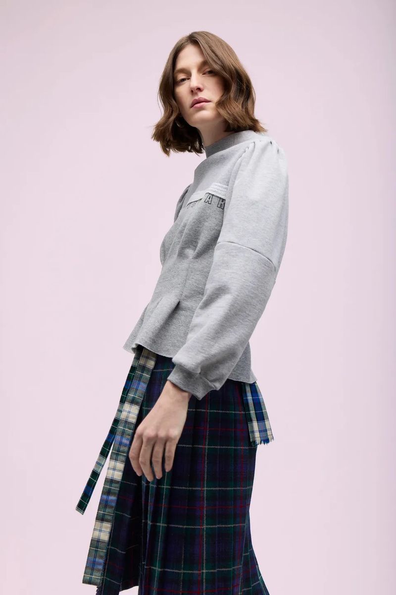 Model Jennae Quisenberry wearing a grey structured sweatshirt and a navy and green tartan pleated skirt against a pale lilac background.