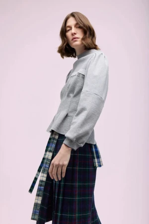 Model Jennae Quisenberry wearing a grey structured sweatshirt and a navy and green tartan pleated skirt against a pale lilac background.