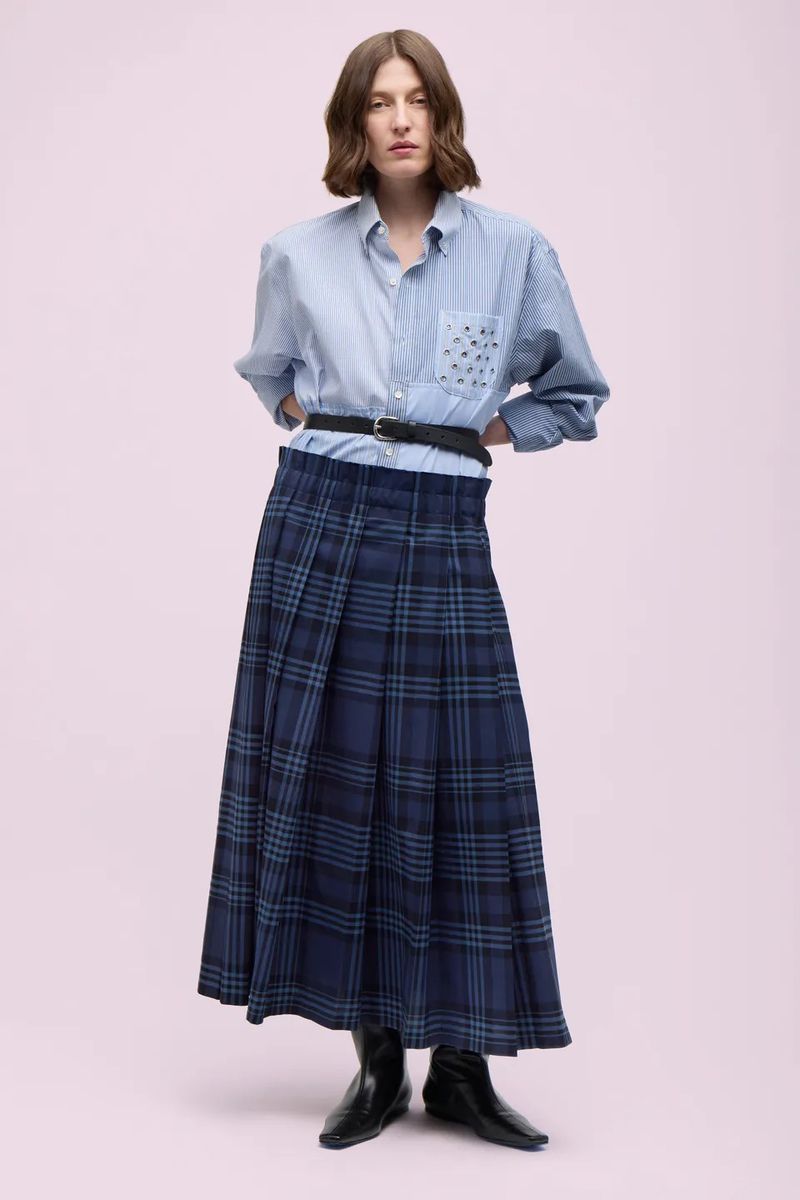 Full-length portrait of an adult woman in an unbuttoned blue striped shirt, plaid skirt, black belt, and black boots against light pink.