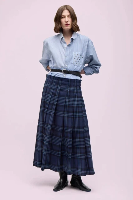 Full-length portrait of an adult woman in an unbuttoned blue striped shirt, plaid skirt, black belt, and black boots against light pink.