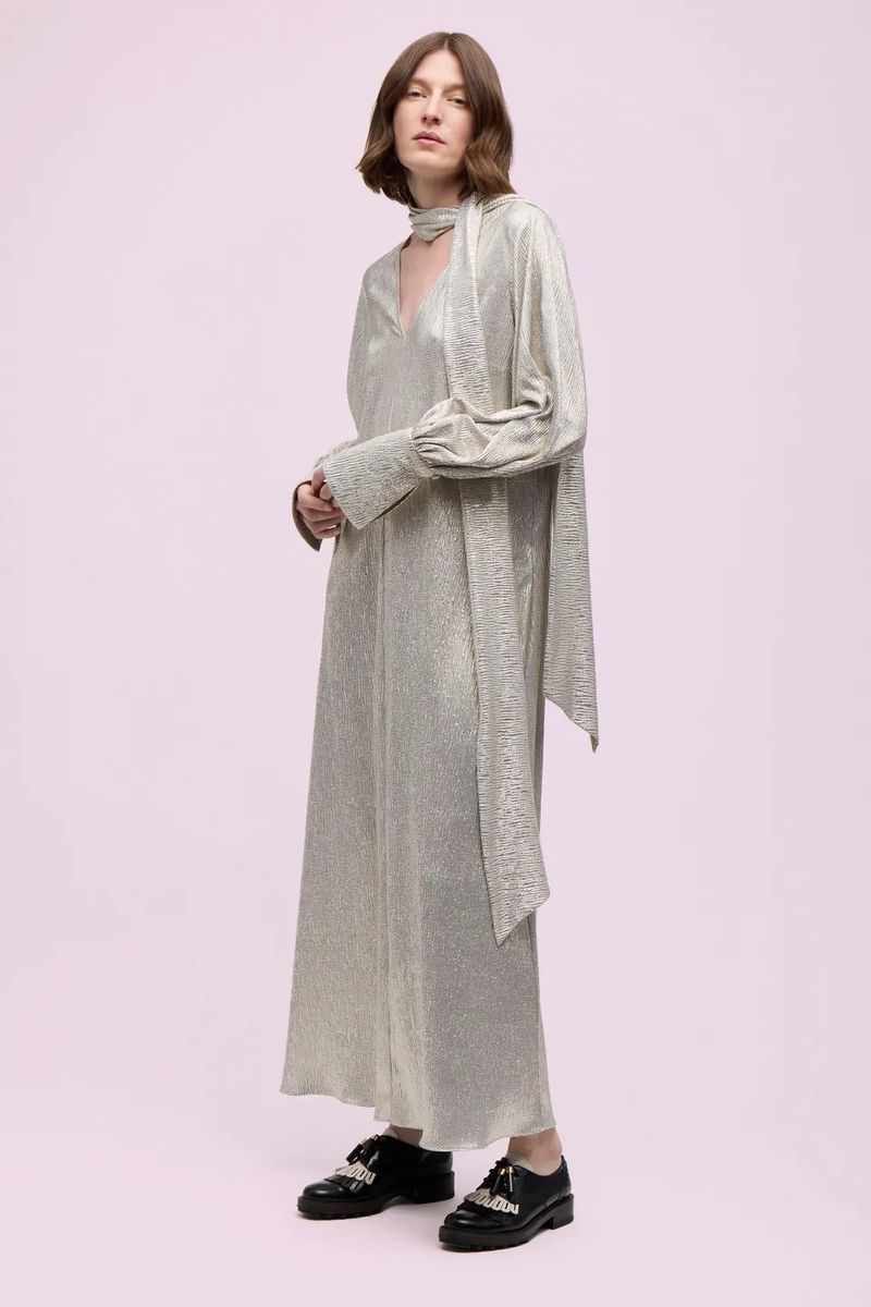 An adult woman is standing in a light pink studio wearing a long, shimmering gold dress and black, decorative shoes in a full shot.