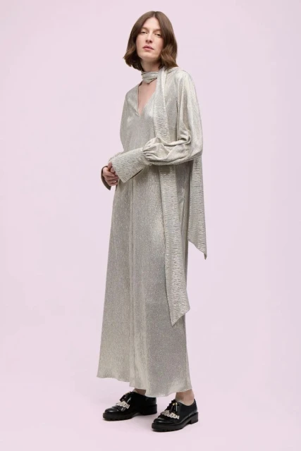 An adult woman is standing in a light pink studio wearing a long, shimmering gold dress and black, decorative shoes in a full shot.