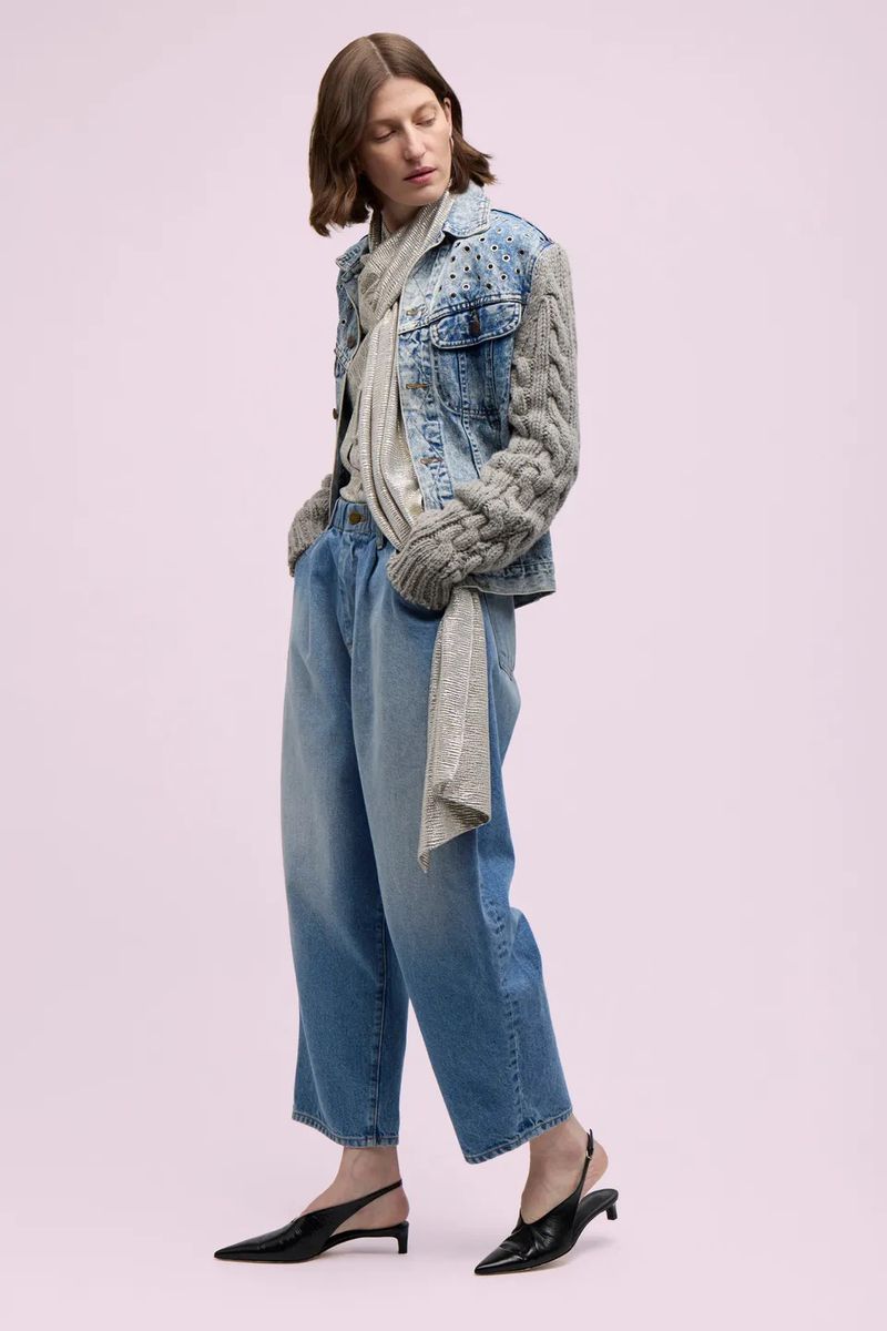 A woman in a denim jacket with knit sleeves, wide-leg jeans, and slingback heels poses against a pale pink backdrop in a studio shot.