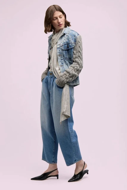 A woman in a denim jacket with knit sleeves, wide-leg jeans, and slingback heels poses against a pale pink backdrop in a studio shot.