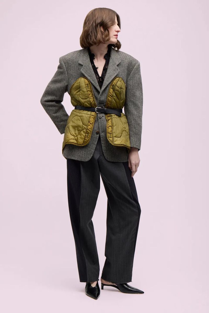 Fashion model wearing a gray tweed blazer with olive green quilted panels, wide-leg trousers, and black heeled mules against a pink background.