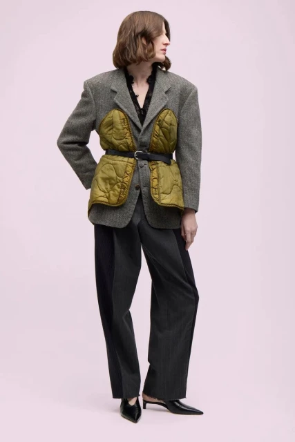 Fashion model wearing a gray tweed blazer with olive green quilted panels, wide-leg trousers, and black heeled mules against a pink background.