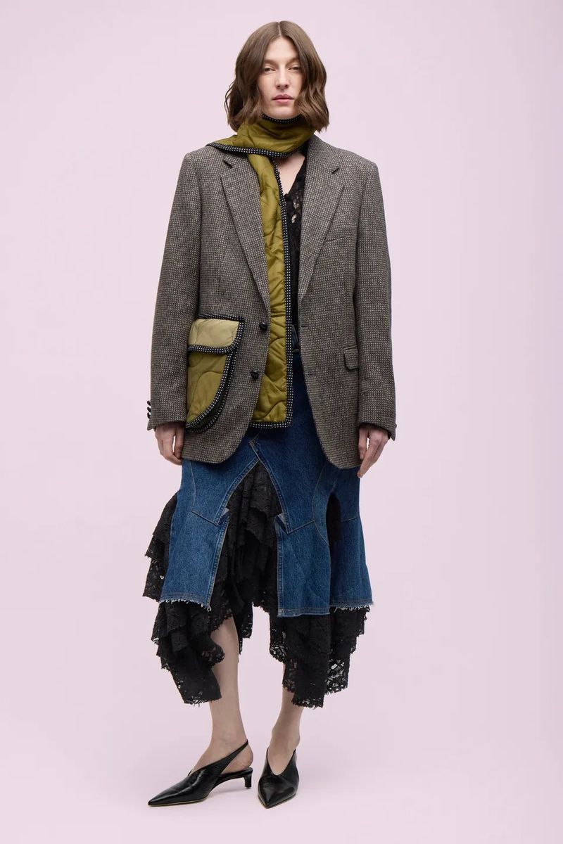 Full-length studio shot of a woman in a grey blazer, olive scarf, and denim skirt with lace. Fashion editorial with a light pink backdrop.