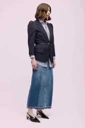 Model wearing a pinstripe blazer, layered shirt, denim maxi skirt, and black slingback heels against a pale backdrop