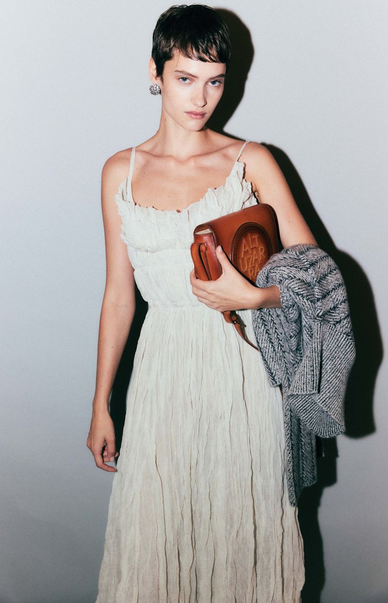 A woman in a white dress poses indoors, carrying a brown leather purse and grey jacket, fashion shoot, portrait, beauty shot.