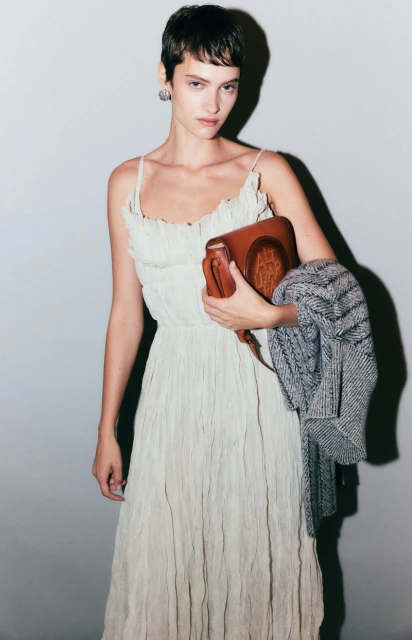 A woman in a white dress poses indoors, carrying a brown leather purse and grey jacket, fashion shoot, portrait, beauty shot.
