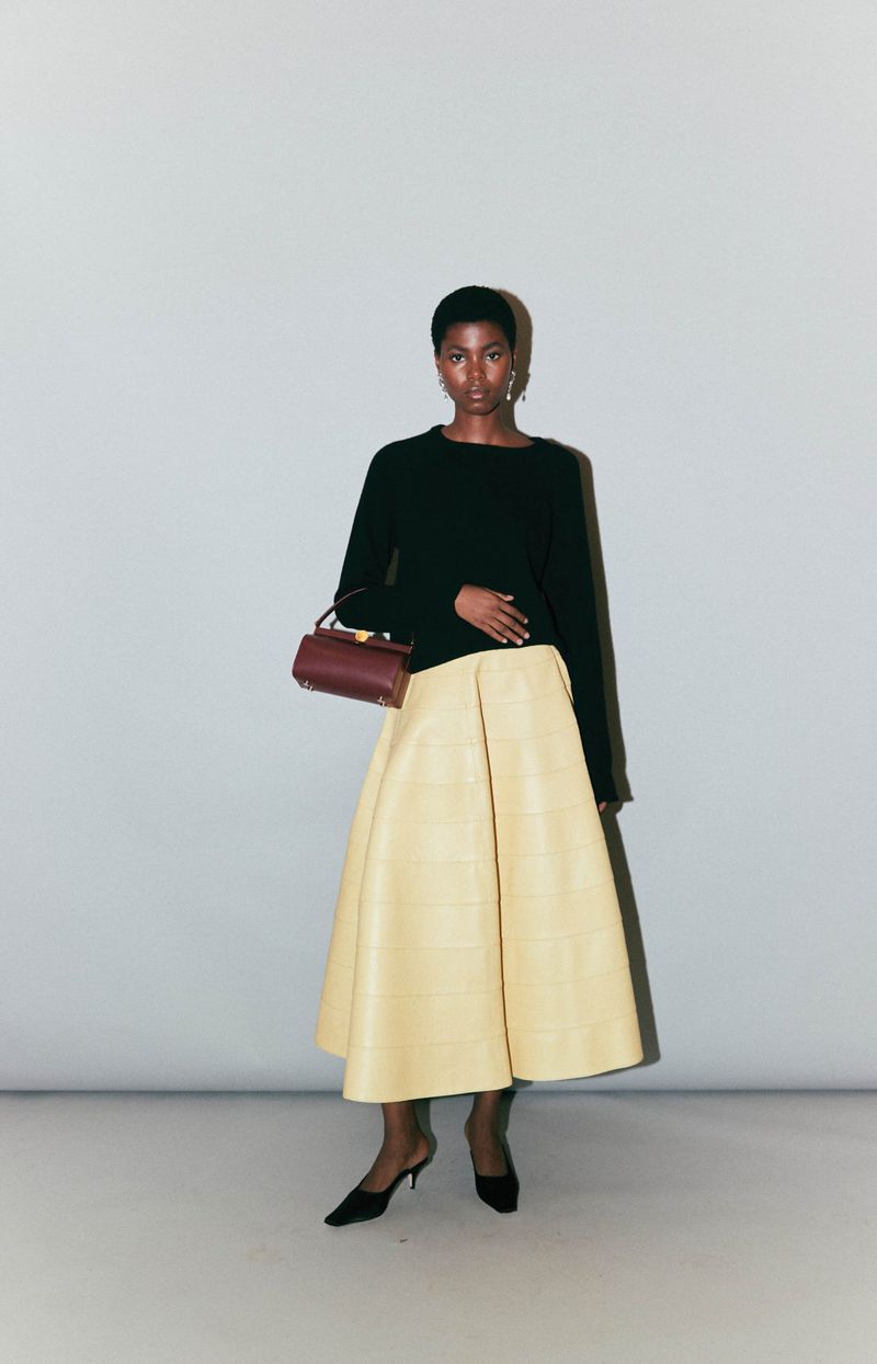 Model wearing a black long-sleeve knit top, pale yellow flared midi skirt, and kitten heel mules with a burgundy top-handle bag.