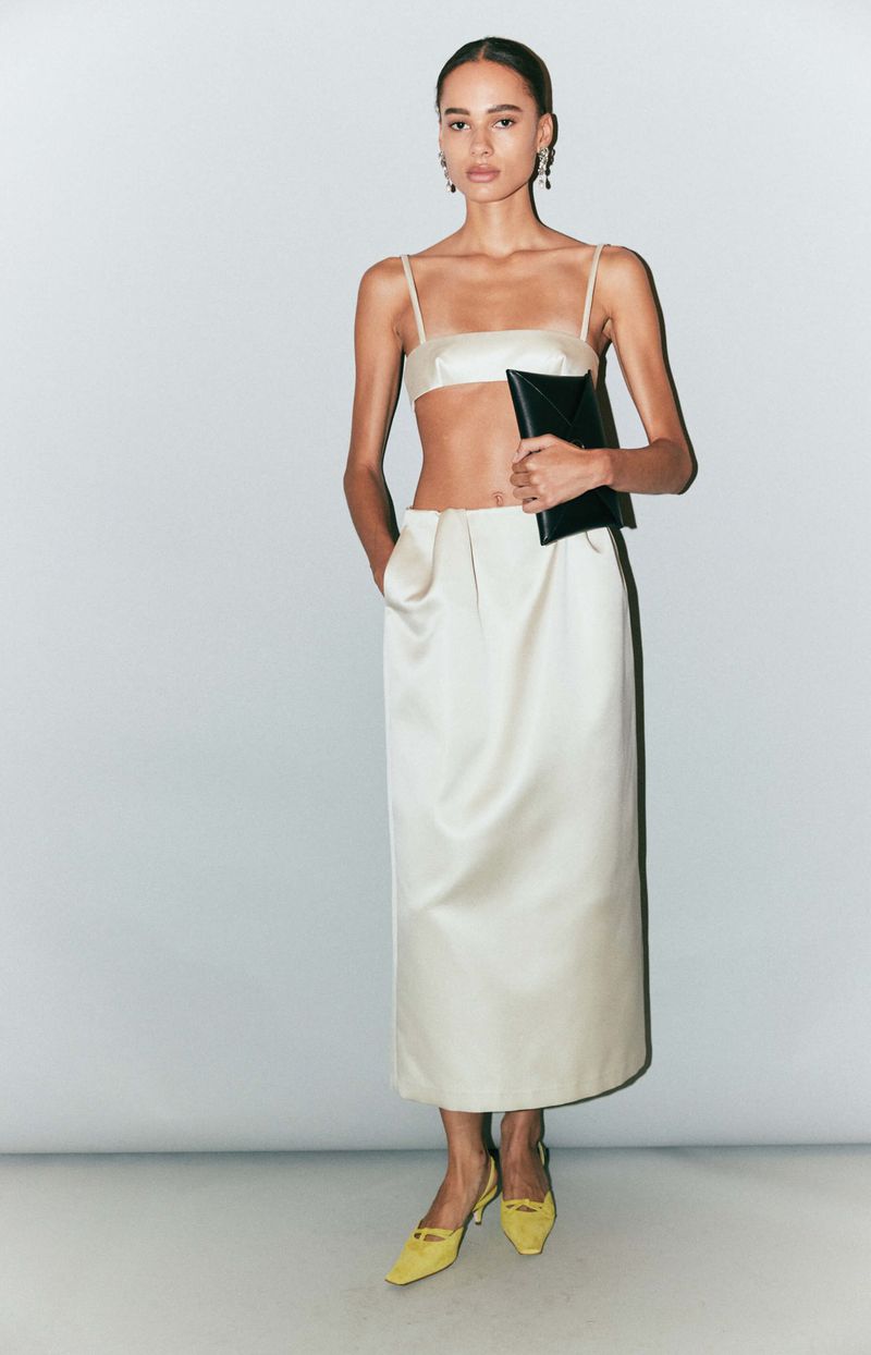 Fashion editorial shot: Woman poses in a cream-colored two-piece outfit and yellow heels, holding a black clutch against a light background.