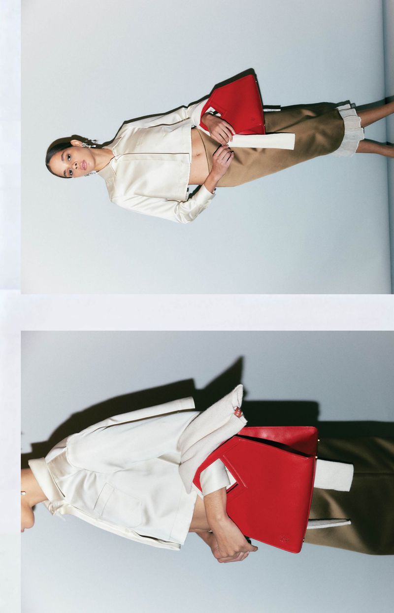 Woman in a light jacket and tan skirt holds a red document case in a fashion shot against a plain light background.