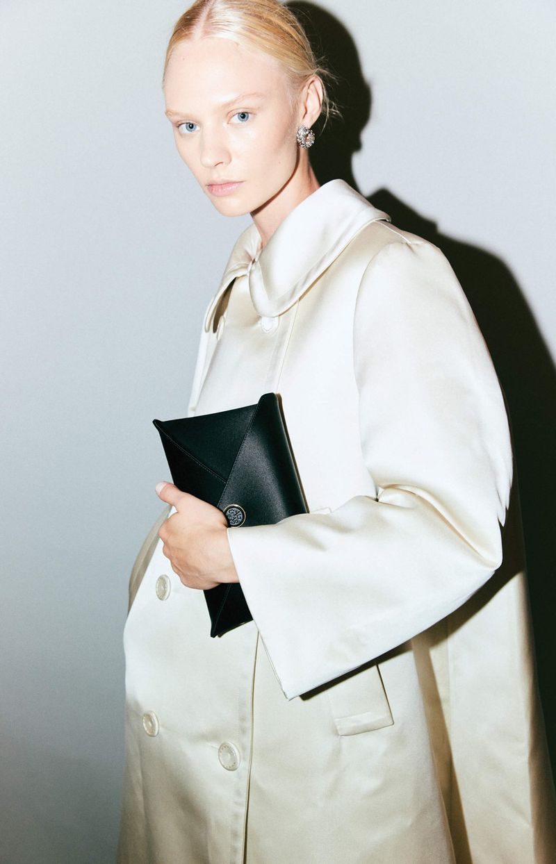 Elegant woman in a satin coat poses indoors holding a black clutch bag, featuring a clean and modern fashion aesthetic.