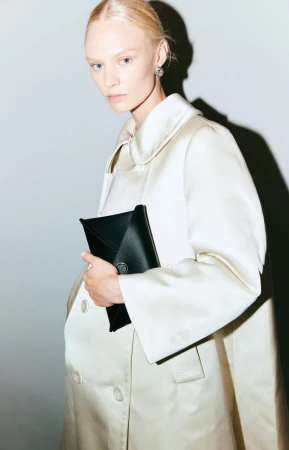 Elegant woman in a satin coat poses indoors holding a black clutch bag, featuring a clean and modern fashion aesthetic.
