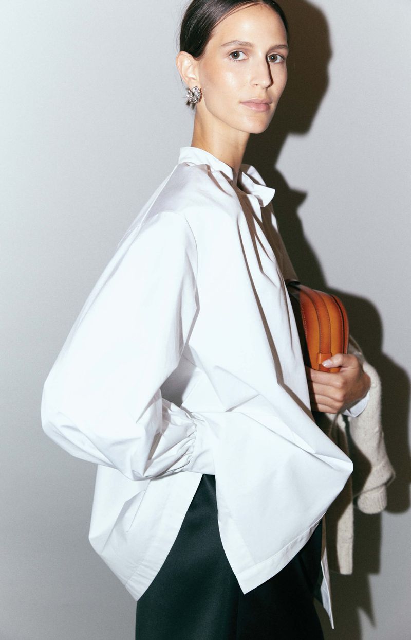 Adult female model wearing a white blouse and black skirt carrying a brown bag, posing in a studio with a white background.