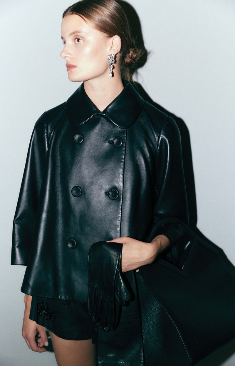 Woman wearing a black leather jacket, shorts, and carrying a large black leather tote bag in a studio setting with bright lighting.