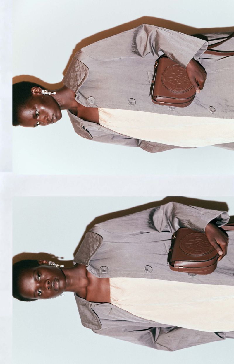 Fashion shot of a Black female model wearing a gray coat and holding a small brown leather bag with a repeating circular pattern.