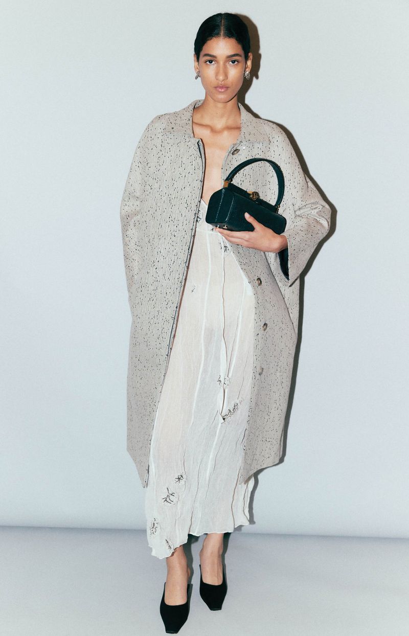Full-length shot of a young adult fashion model wearing an overcoat, a dress, and black heels, holding a small black purse. 