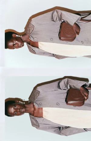 Model wearing a grey coat and holding a brown leather handbag against a plain background