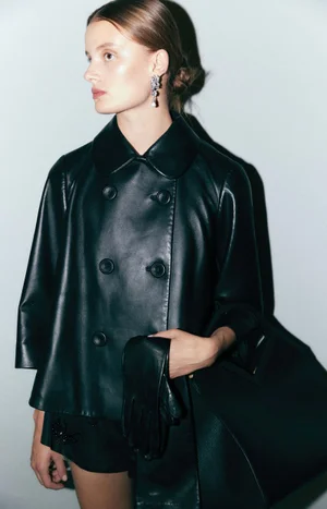 Model wearing a black leather double-breasted cape jacket and gloves while holding a large matching black tote bag against a neutral backdrop.