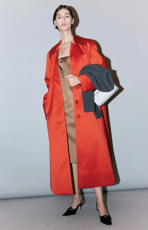 Model in a bright red trench coat holding a grey sweater and white handbag against a plain grey studio backdrop.