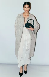 Model wearing a speckled grey coat and white dress, holding a dark handbag against a neutral studio backdrop